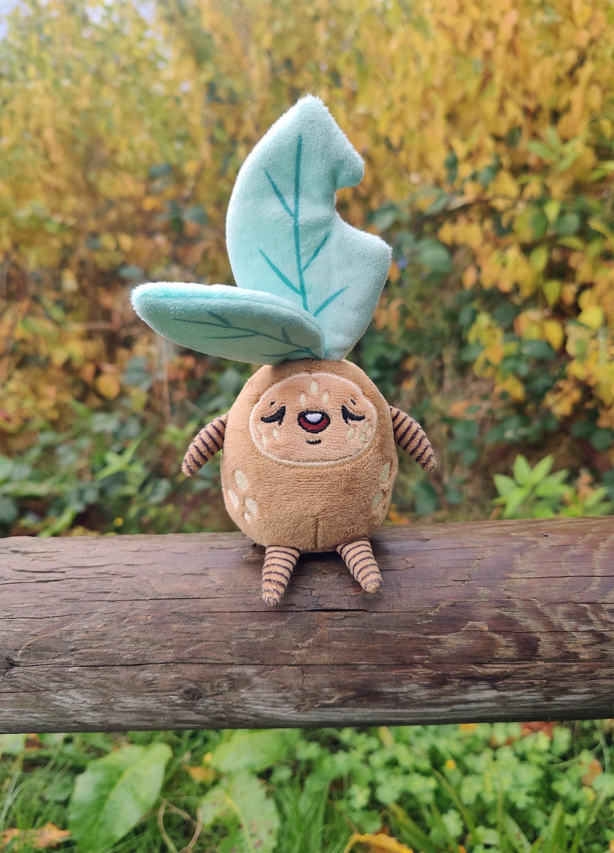 Plush toy resembling a sleepy leafy plant creature sitting on a log with a natural background