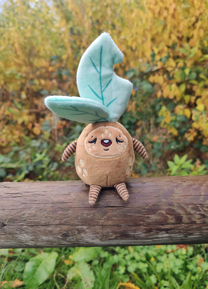 Plush toy resembling a sleepy leafy plant creature sitting on a log with a natural background